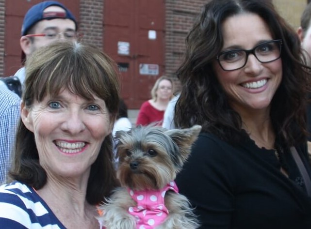 Elaine aka Julia Louis-Dreyfus in big black glasses poses for a picture with a smiling Elly and her dog, Schmitty The Weather Dog, a Yorkshire terrier wearing a pink polka dot dress.