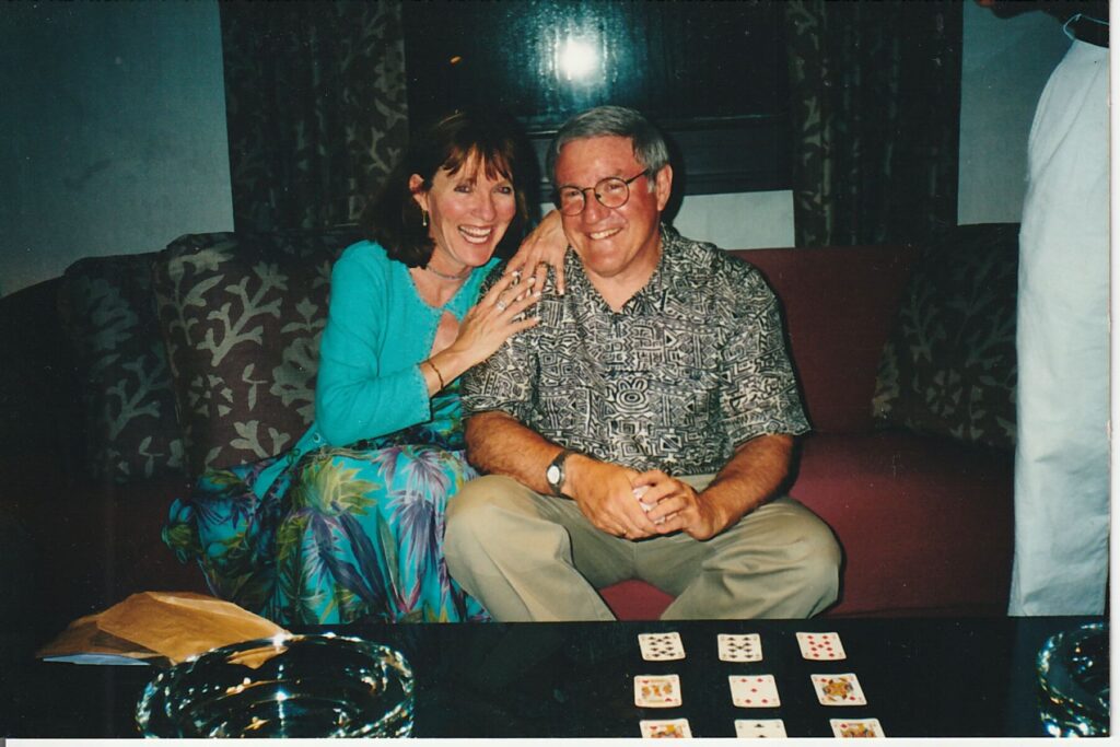 Elly McGuire, host of her podcast, Shaking The Bushes, poses with Dr. John Eckstein from the Mayo Clinic. They are sitting on a couch in the lounge of a yacht in Greece in 2001.