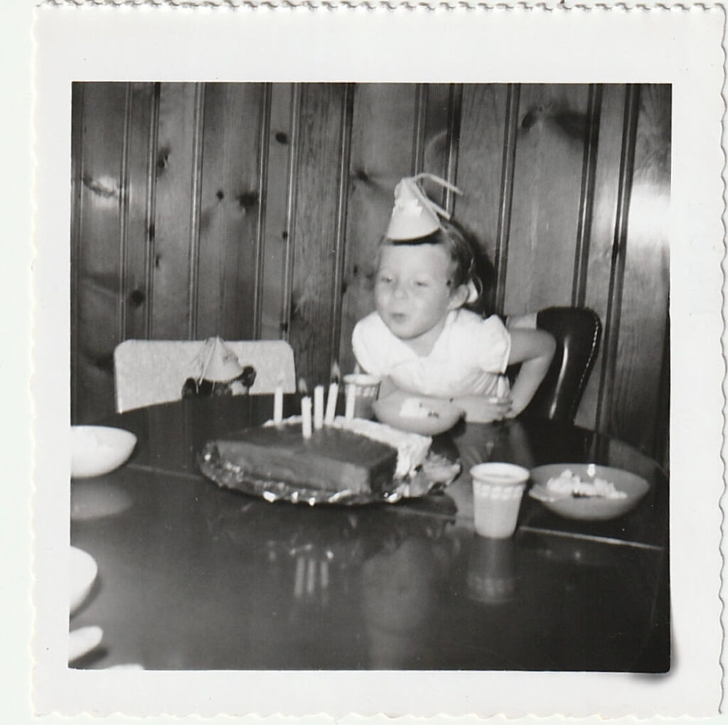 A vintage picture of Podcast host, Elly McGuire, at age 4, wearing a white dress and birthday hat- blowing out the candles of her birthday cake.