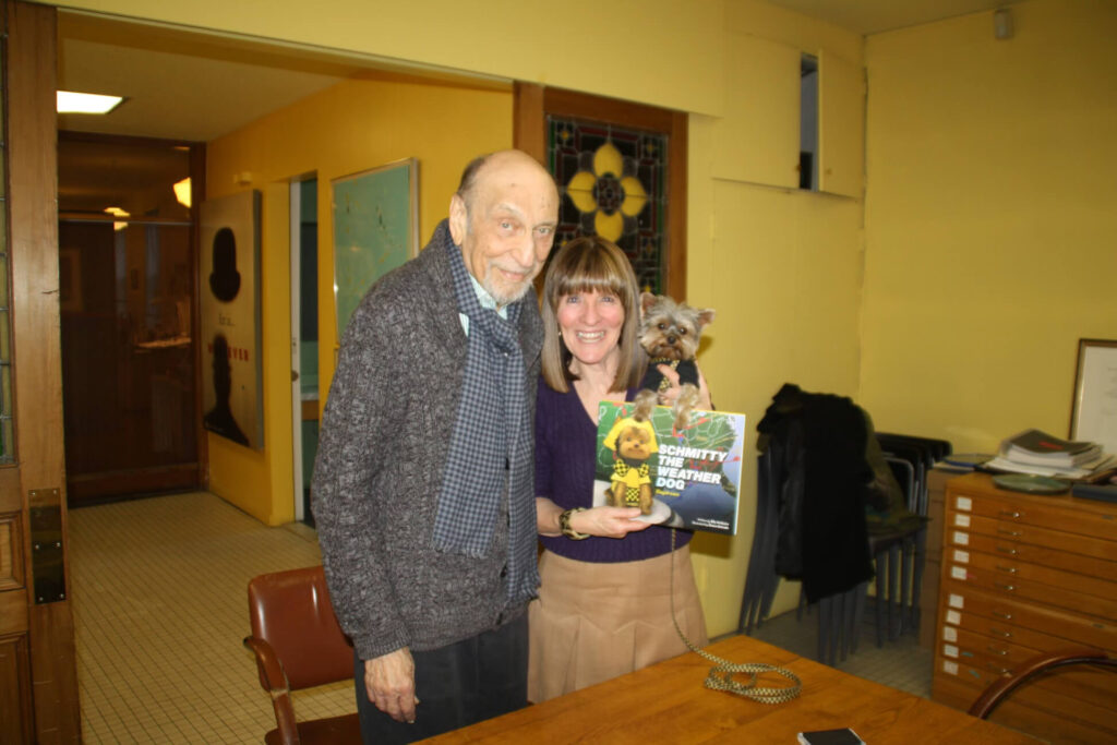 A picture of Elly McGuire holding up her Schmitty The Weather Dog: Daydream children's book with the real live Schmitty The Weather Dog ( s five pound Yorkie) leaning on the book. Together they are standing with the famous graphic designer, Milton Glaser.